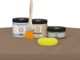 Retique It Barn Wood Kit with Matte Tripletique, Light Wood Wood'n Primer, Barn Wood Gel Stain Alternative, paintbrush, and yellow sponge on a tabletop.